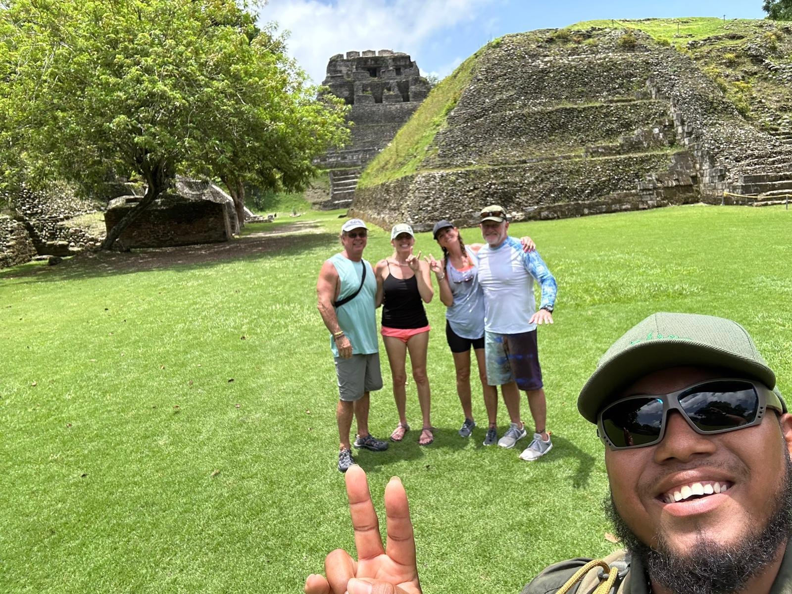 Four guests smiling with their guide in front of an ancient Maya pyramid, the guide holding the camera for a selfie