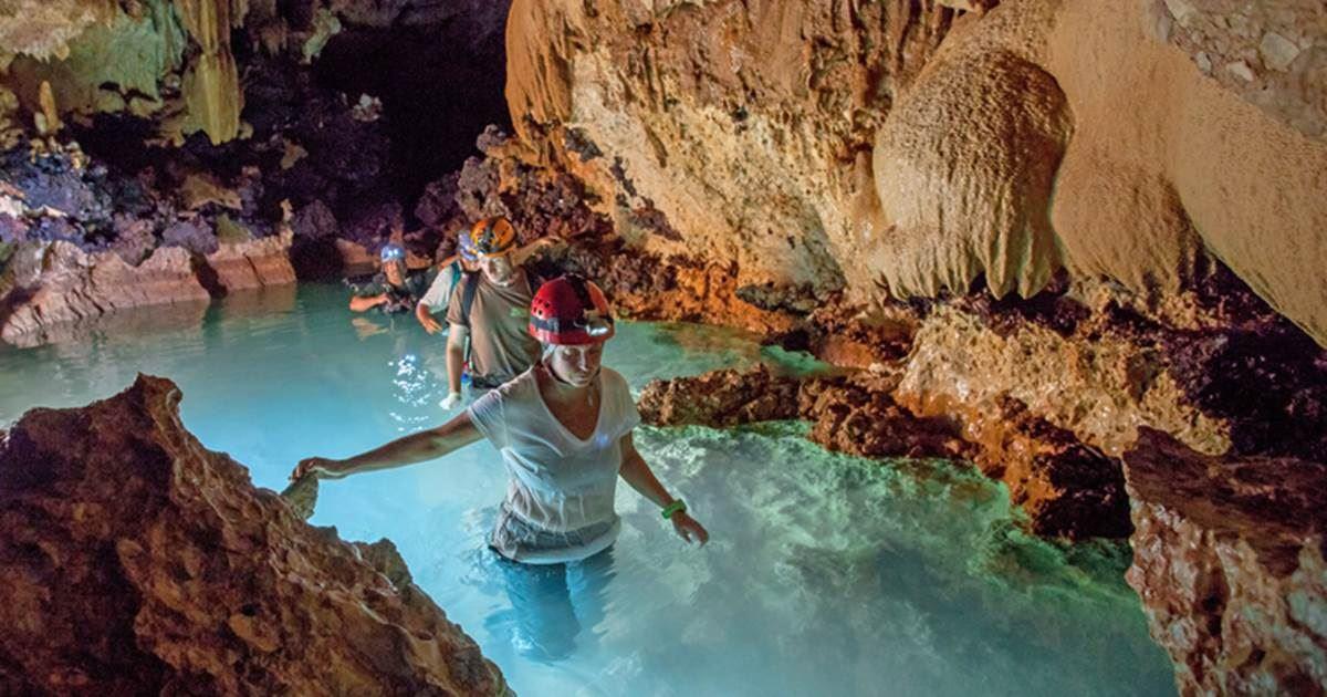 A guest in a red helmet wading through a glowing turquoise cave pool on a guided adventure