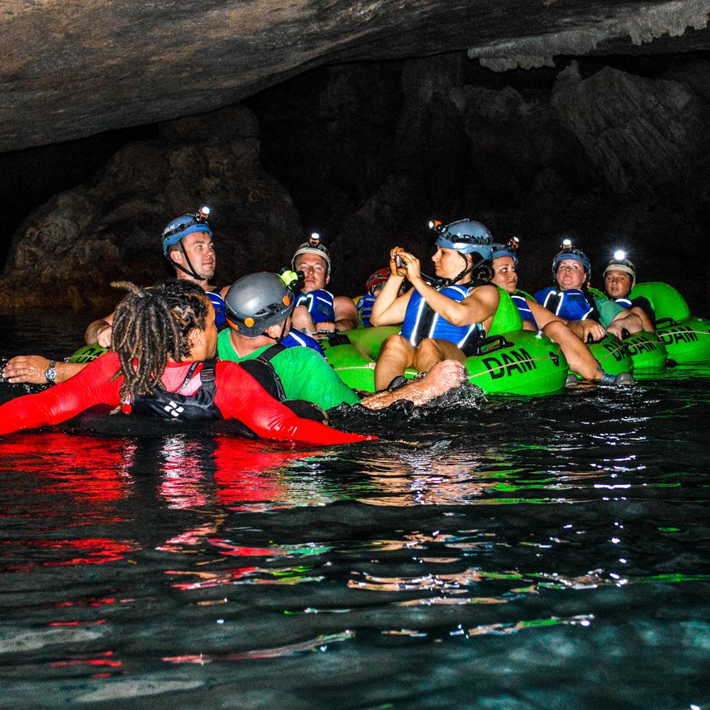 A group of guests in helmets with headlamps floating on green tubes through a dark cave, their guide beside them