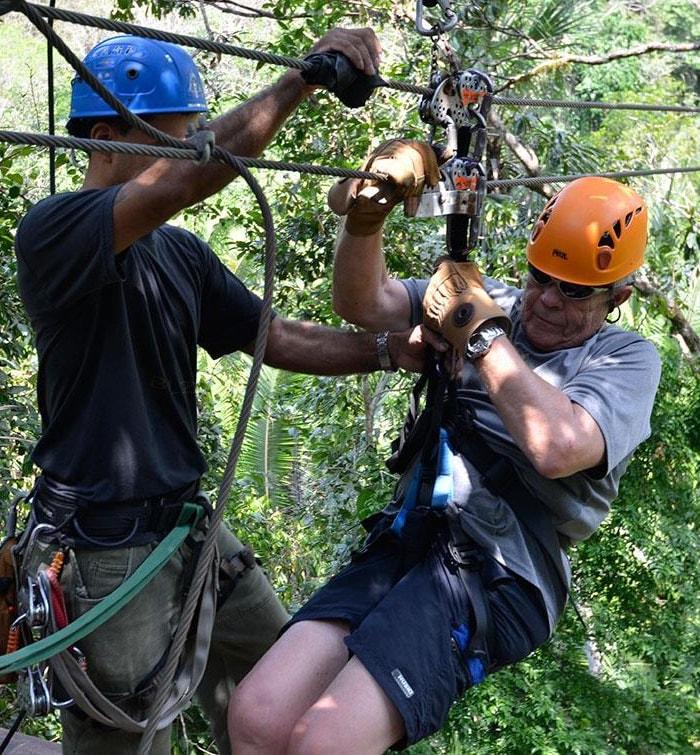 A guide helping a guest in an orange helmet clip onto a zipline cable high in the forest canopy