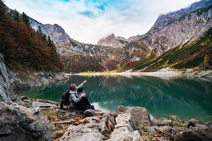 Two travelers sitting side by side on the rocky shore of a turquoise mountain lake, one pointing toward the peaks ahead