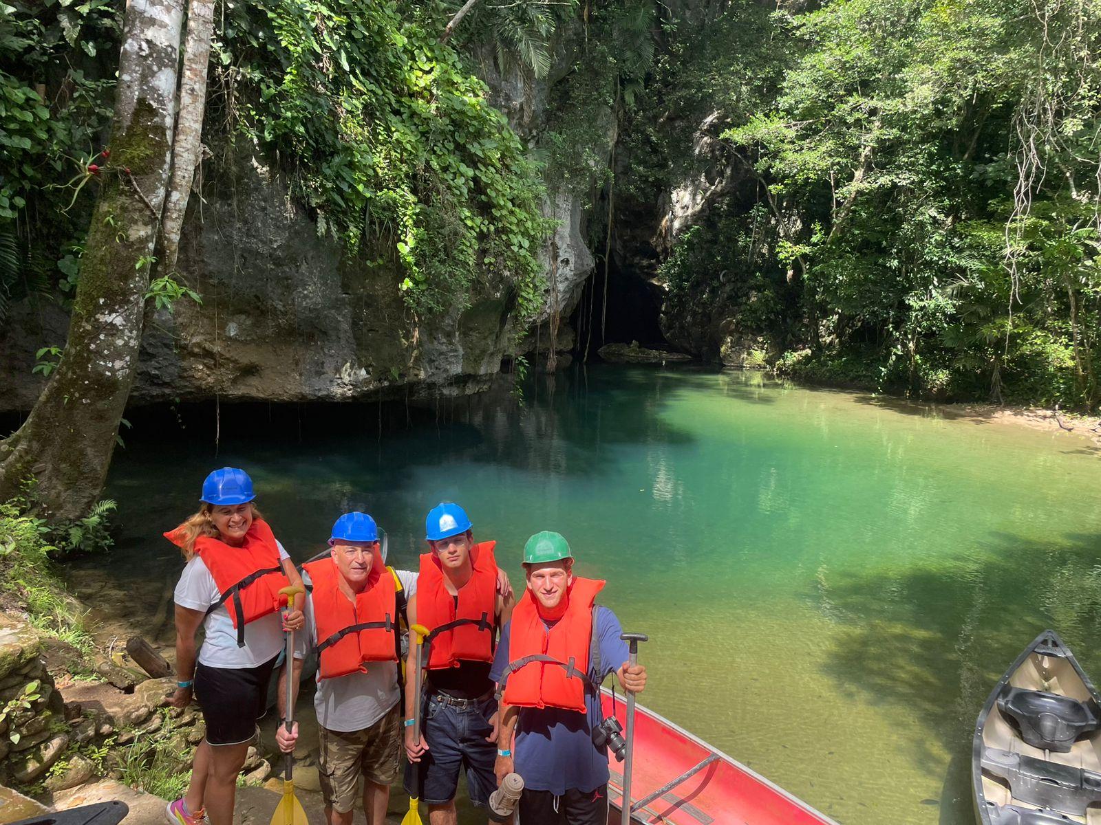 Four guests in hard hats and orange life jackets holding paddles beside a canoe on turquoise water at the entrance to Barton Creek Cave in Belize
