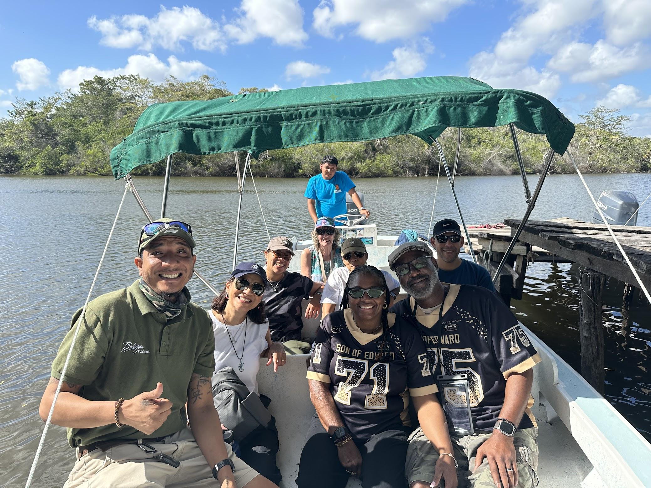 A Belize Fun Tours guide in a green shirt gives a thumbs-up beside smiling guests on a boat under a green canopy on the New River, a wooden pier behind them