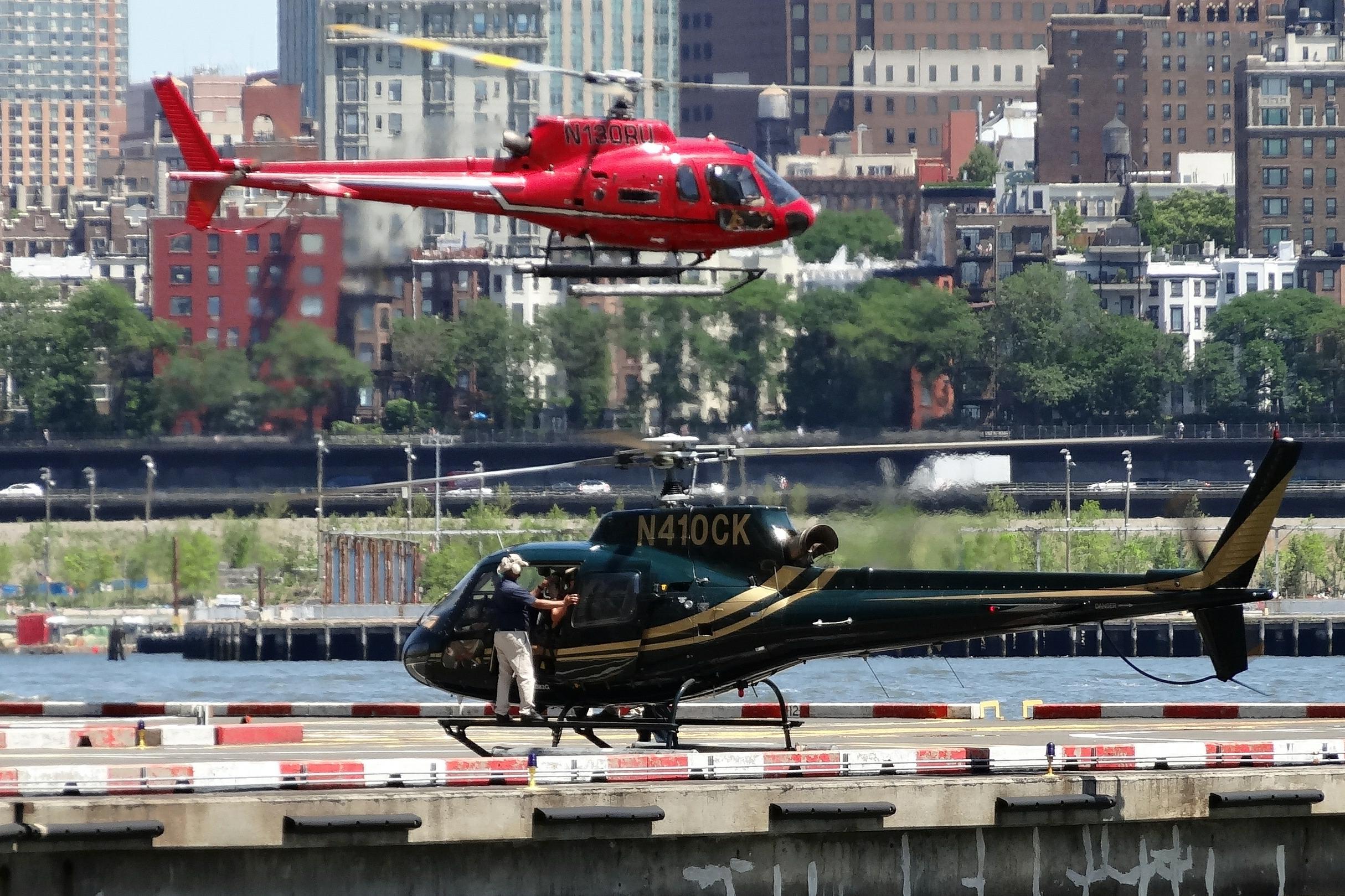 A crew member stands at the open door of a parked tour helicopter on a New York City heliport while a second helicopter hovers overhead, city buildings behind