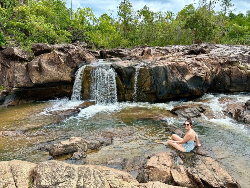 A traveler sitting in a natural rock pool at the foot of a small waterfall, surrounded by tropical forest