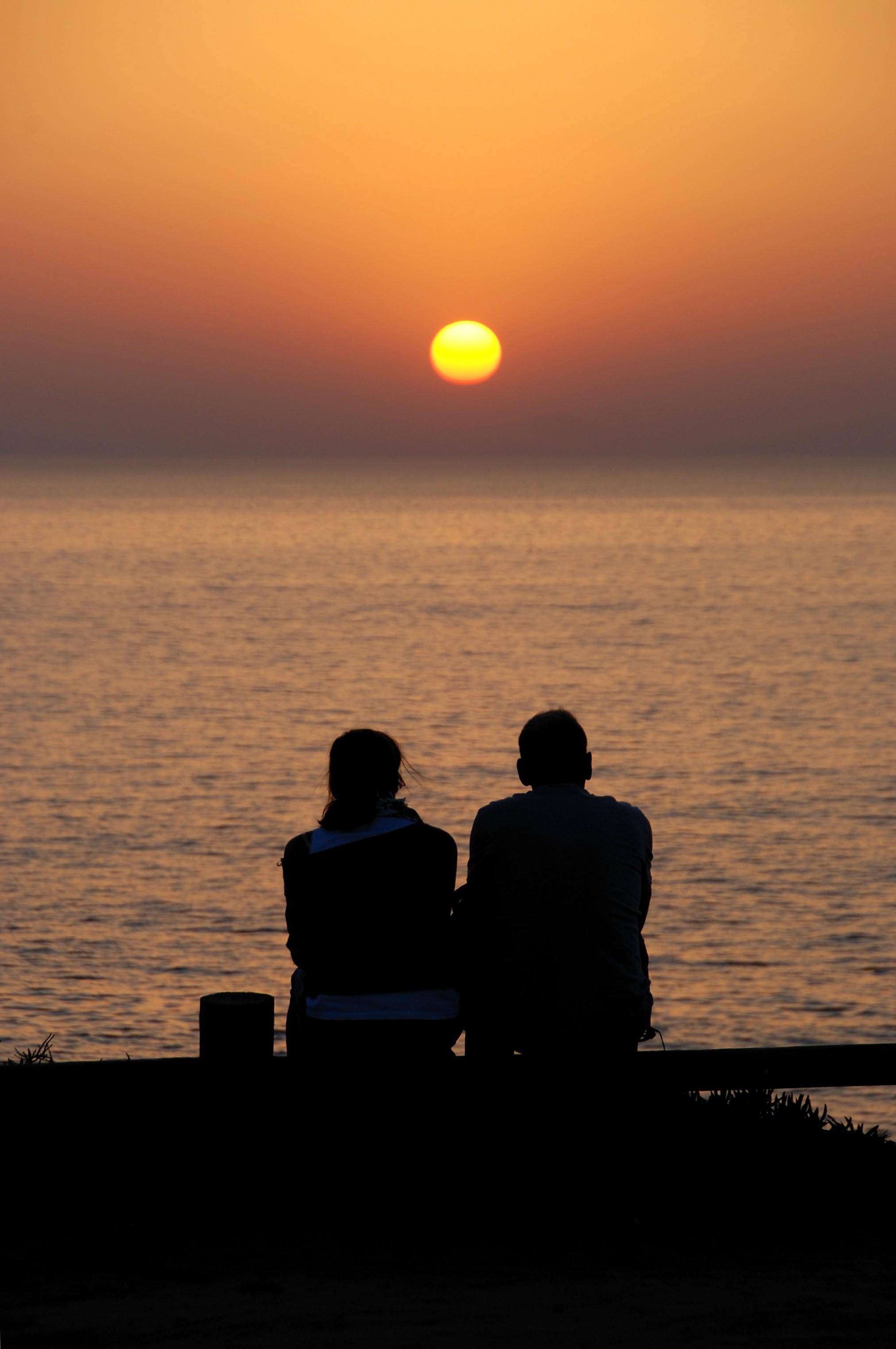 Two people seen from behind, sitting side by side at a seaside rail, silhouetted against a glowing orange sunset over the ocean