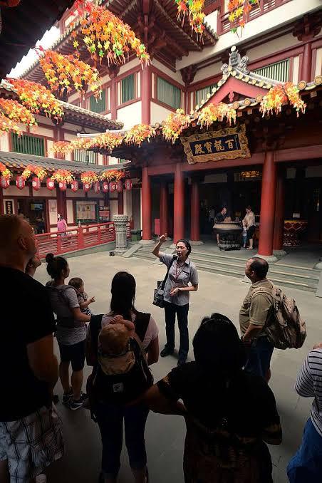 A tour guide addresses a group of visitors in a temple courtyard hung with orange lanterns, red pillars and carved roofs behind her